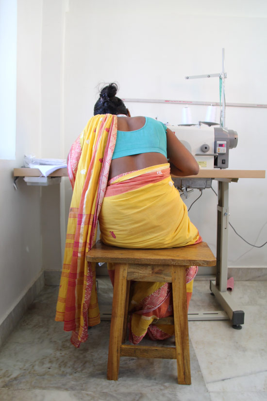 South Asian woman sits at a sowing desk with her back to the camera.